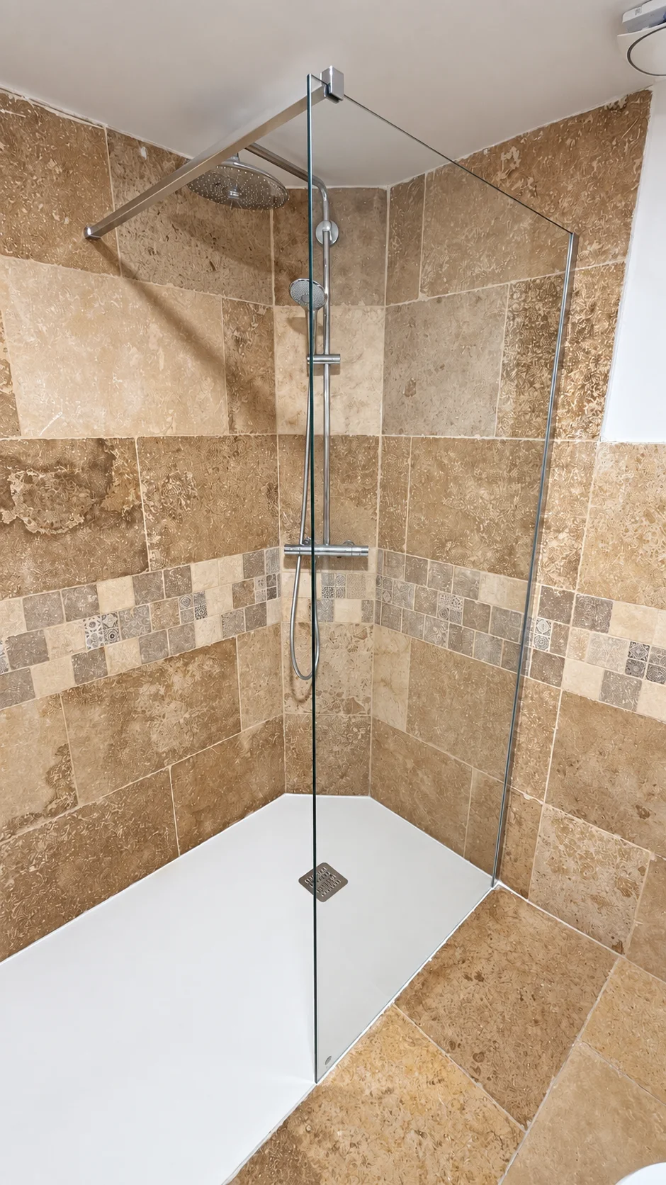 Douche avec receveur blanc, paroi vitrée fixe et colonne chromée sur carrelage beige.