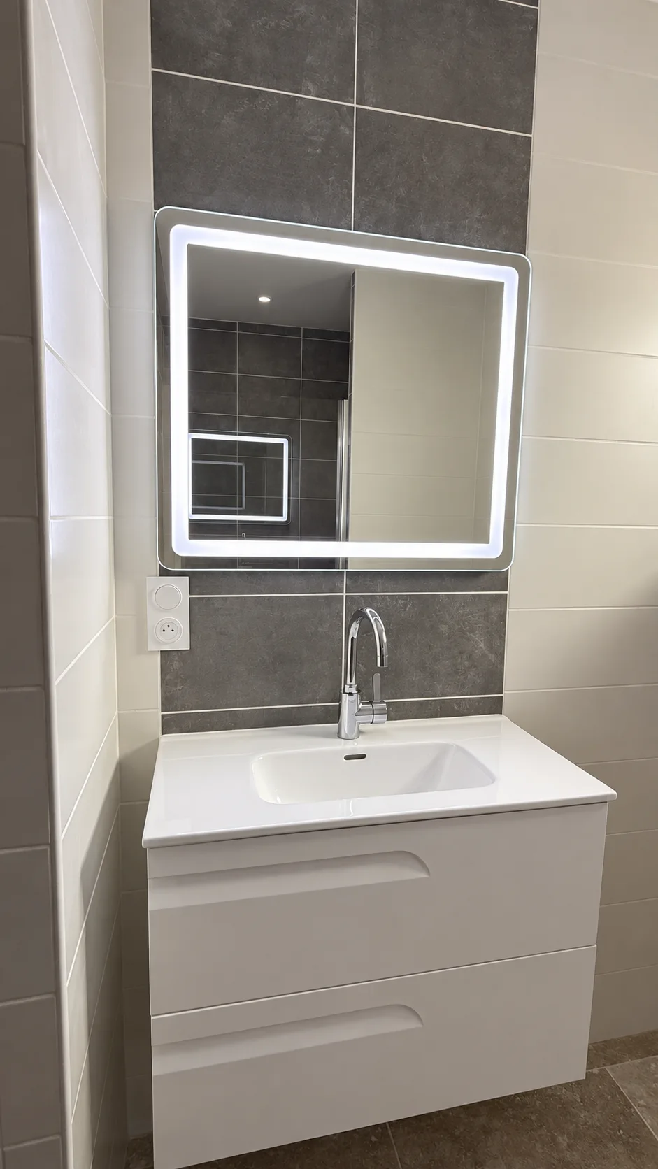 Meuble vasque blanc suspendu sous un miroir à éclairage périphérique, sur carrelage gris et blanc.